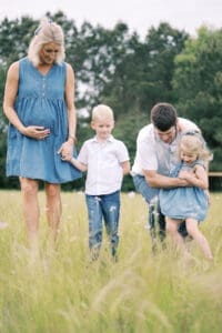 A family of four dressed in denim walks in a field of tall grass and flowers as the father holds his daughter