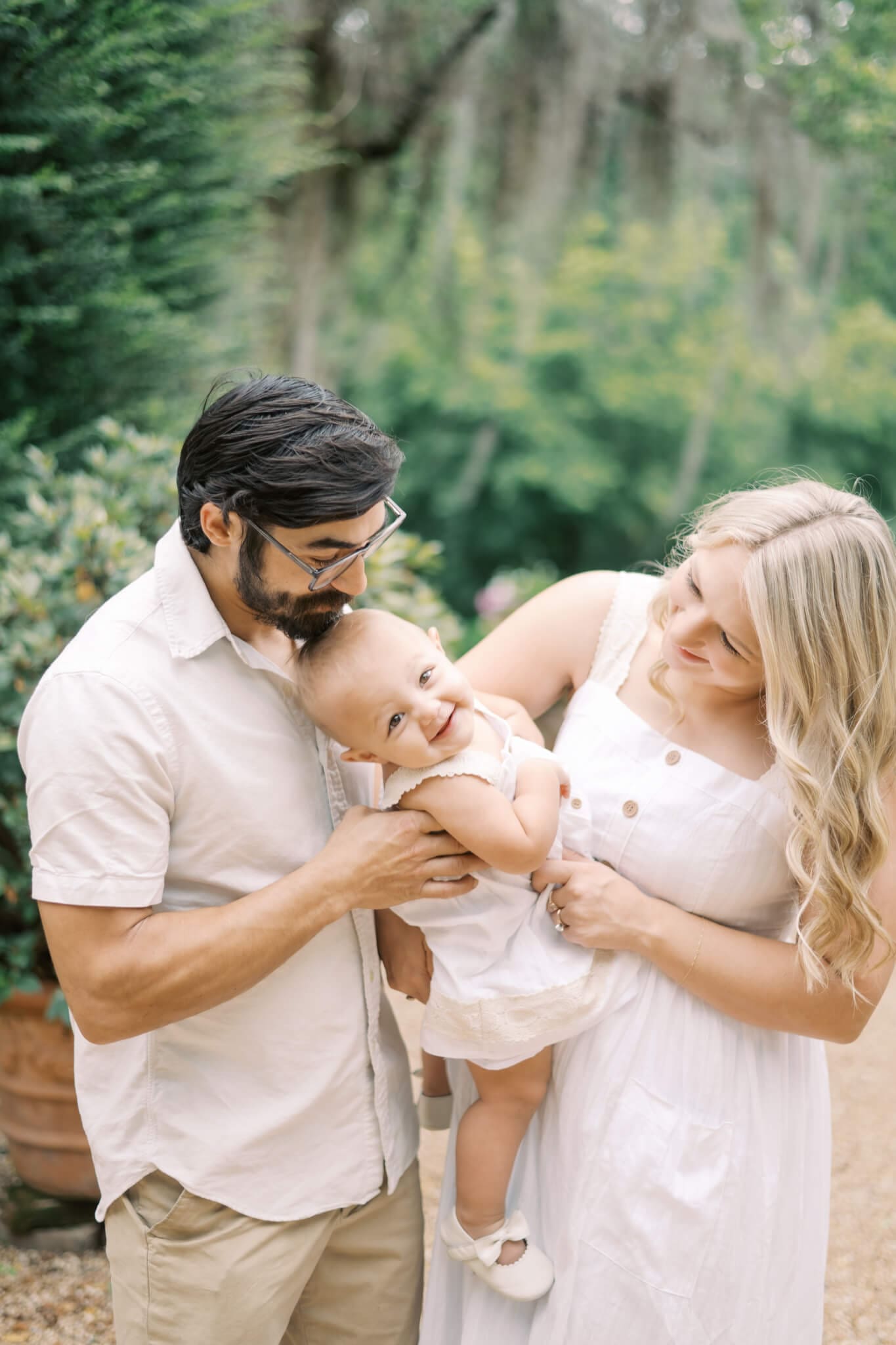 A mom and dad tickle their toddler daughter while she laughs and looks at the camera