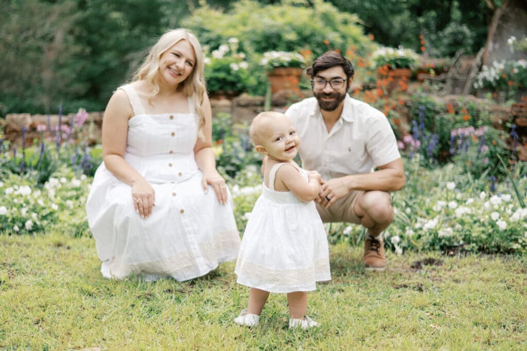 A toddler looks over her shoulder and smiles at the camera while the parents look lovingly at her