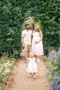 A toddler smiles and walks toward the camera while the parents walk behind her