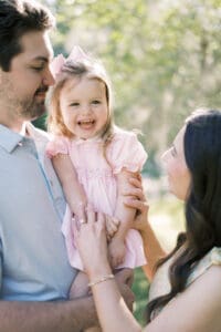 A mother and father hold their toddler in pink while she laughs into the camera
