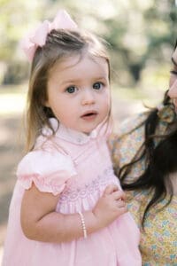 A close up image of a girl wearing a pink dress and bow while she looks at the camera