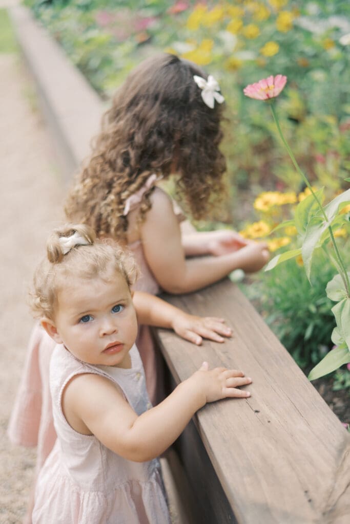 A toddler with blue eyes leans on the side of a raised flower bed while looking at the camera