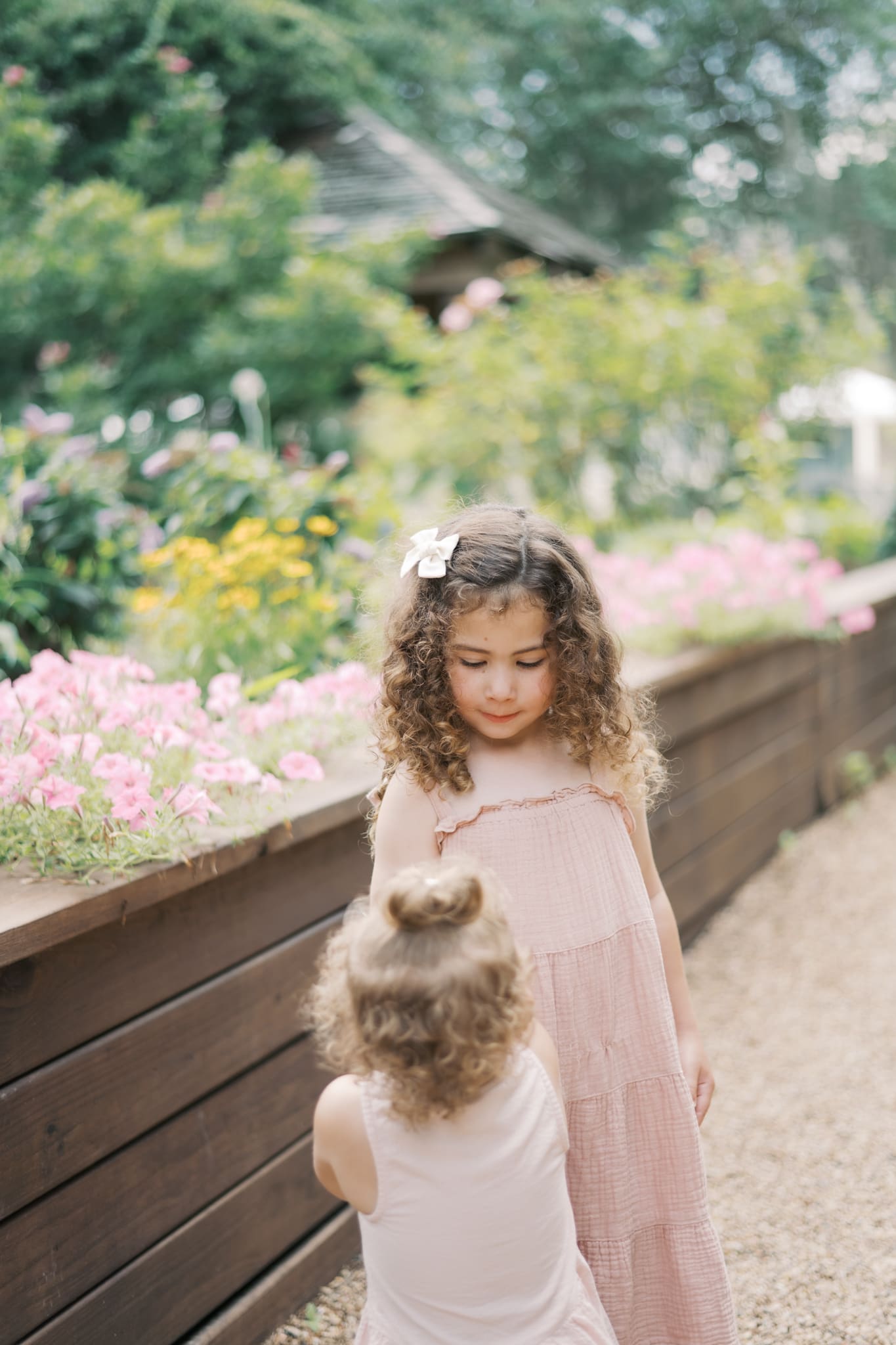 Two sisters in pink dresses and curly hair hold hands while the older sister looks down at the younger sister