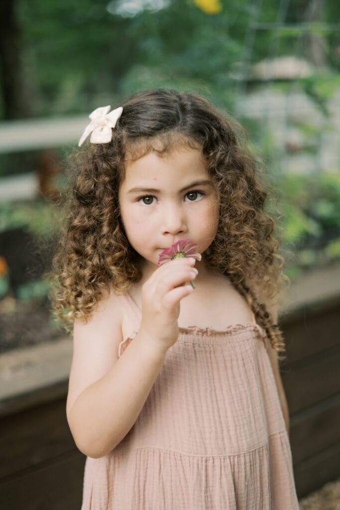 A little girl with curly hair smells a purple flower while looking at the camera
