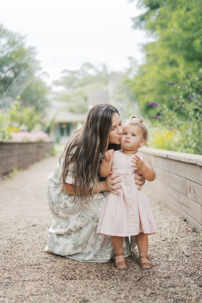 A mother kneels down and gives her toddler a kiss on the check