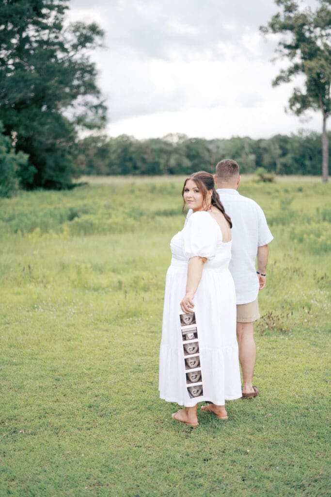 a dad leads the mom by the hand as they walk in a pasture as the mom holds her baby sonogram behind her and as she looks back at the camera