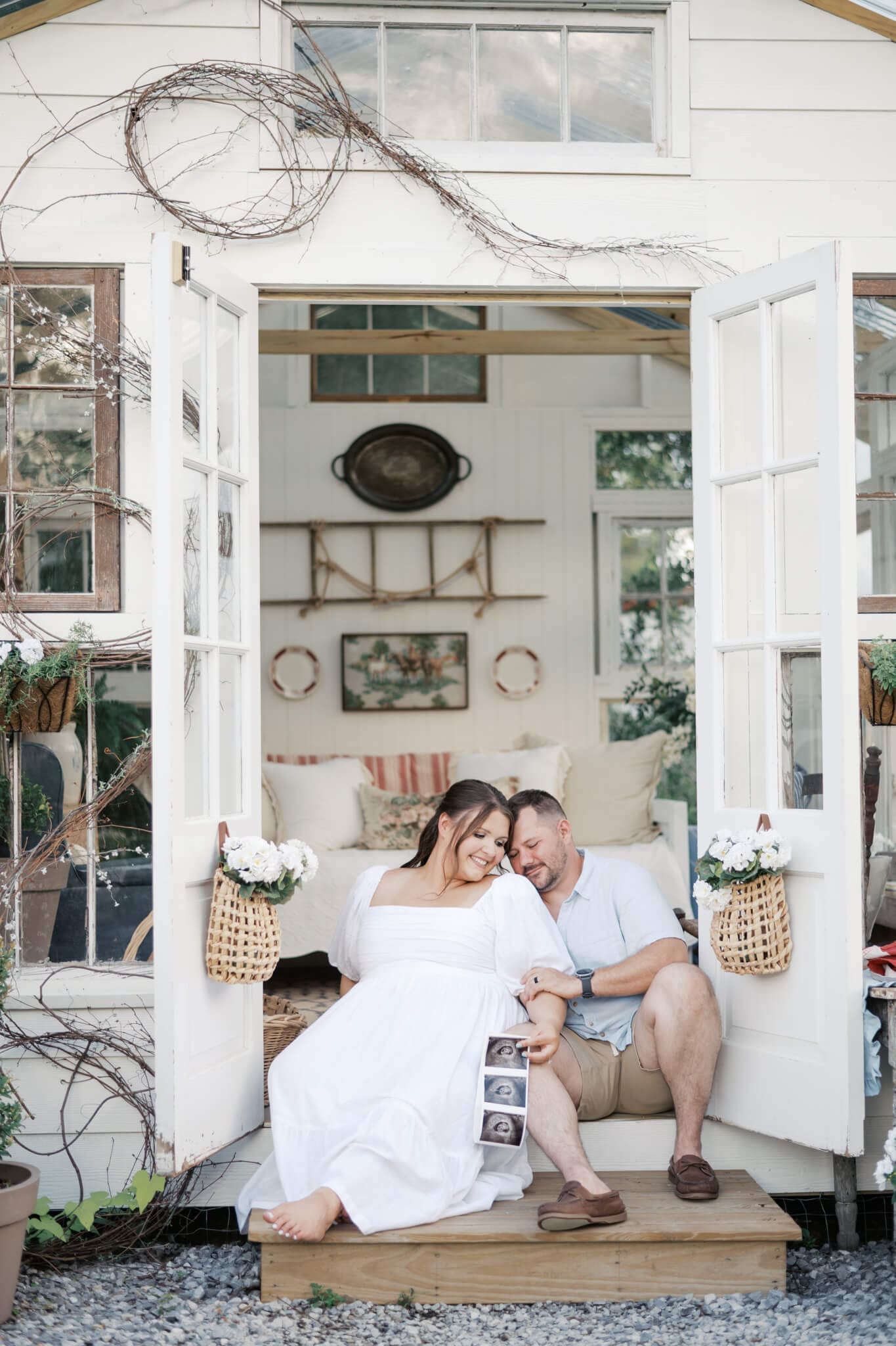 A mom and dad sit on the steps of a farm house as they snuggle close with a baby sonogram