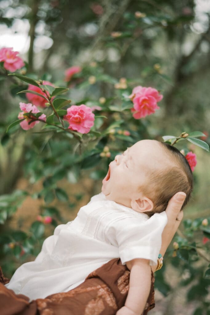 A yawning baby being held by his mother under a camellia tree