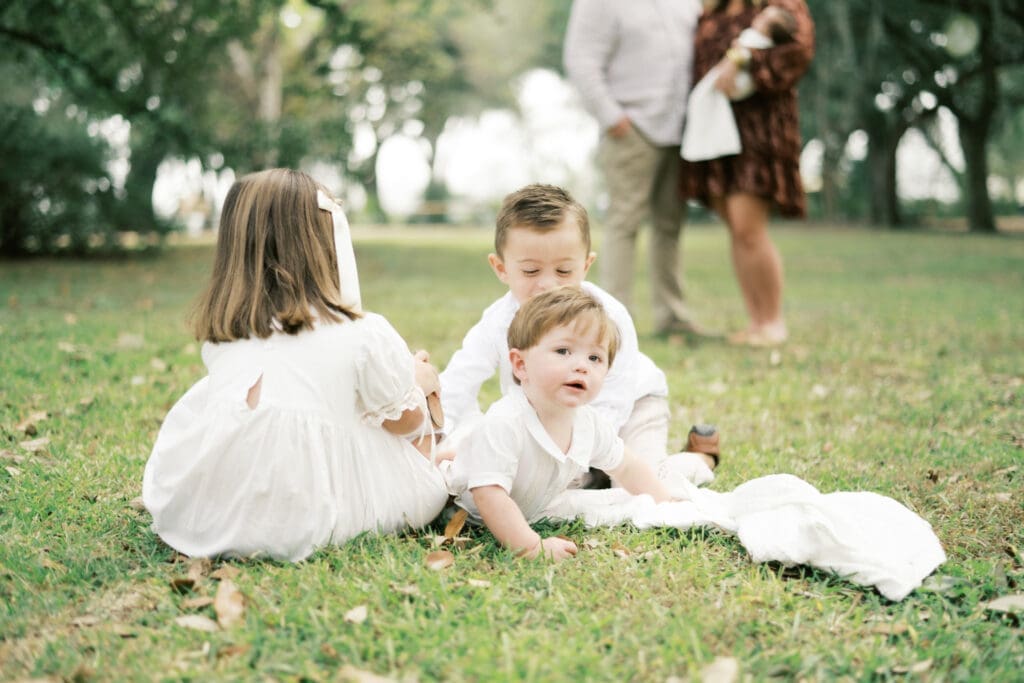 Three siblings play on a white blanket while the youngest brother pauses to look at the camera