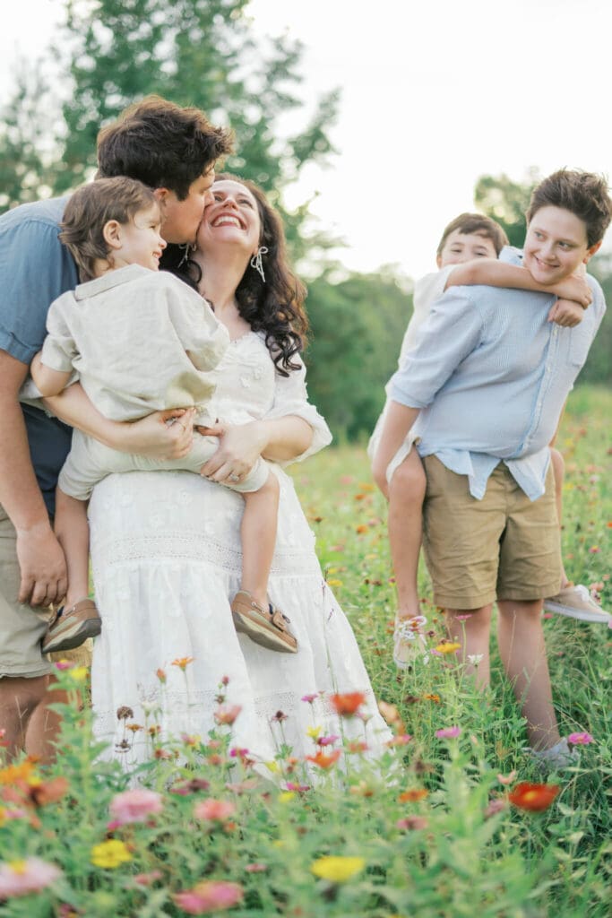 A family of 5 stand in a wildflower field as the dad kisses the mom on the cheek