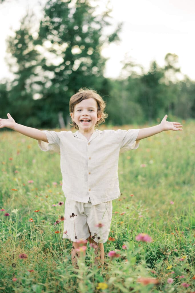 A toddler boy holds his arms out wide as he stands in a field of wildflowers