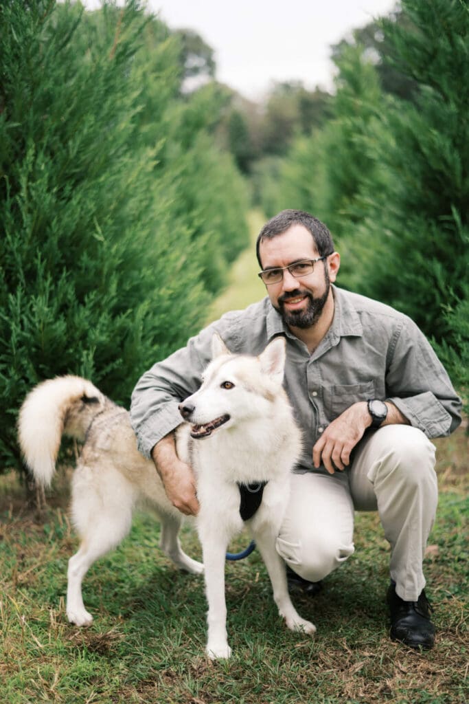 A dog owner kneels next to his white husky dog and smiles at the camera amongst Christmas Trees