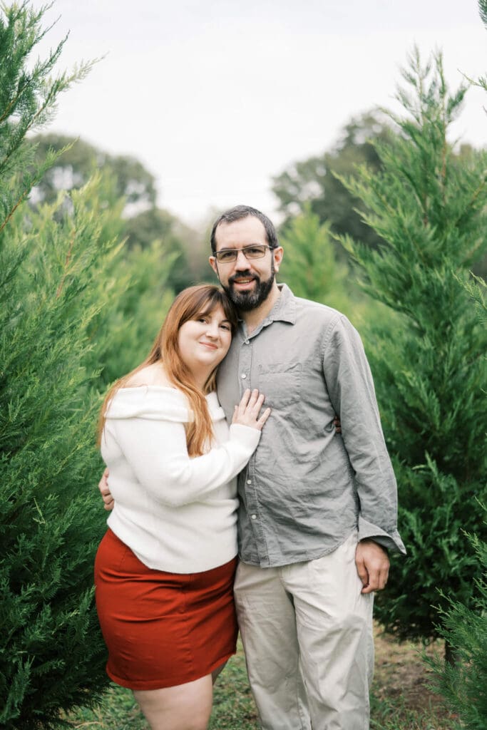 A couple wearing Christmas attire smile at the camera amongst Christmas Trees